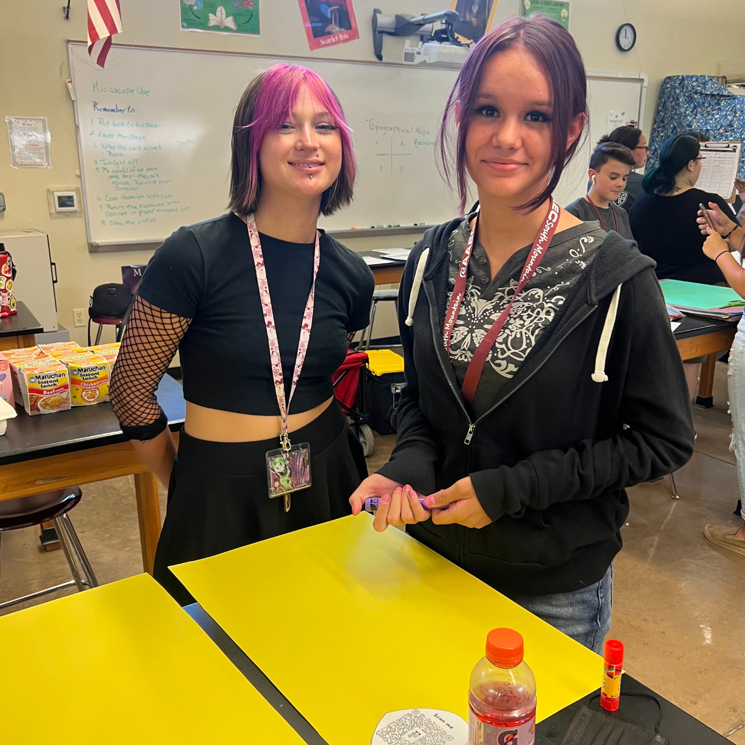 two girls posing in front of a yellow table in a classroom.